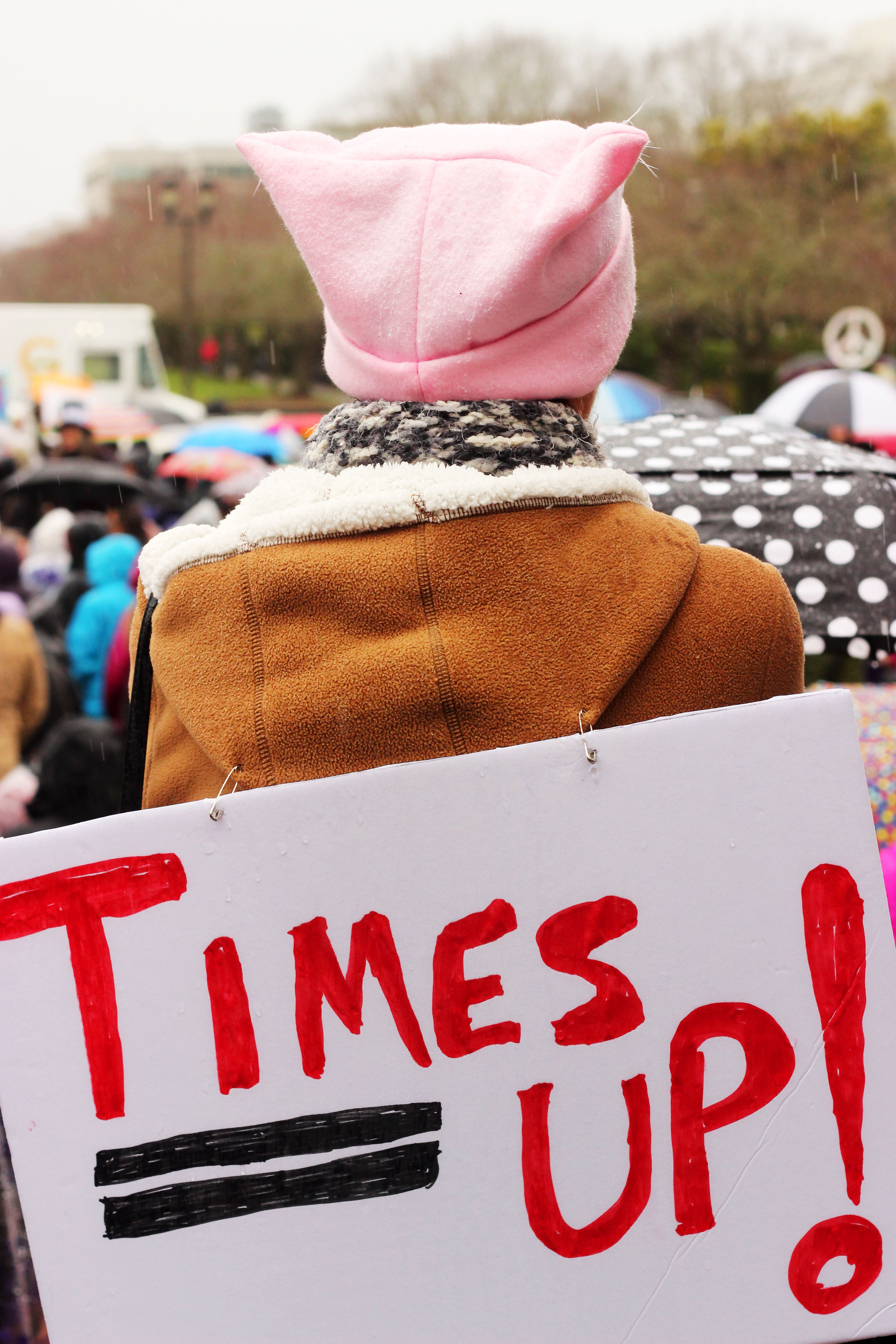 Woman standing in a brown coat wearing a pink hat with a sign on her back saying "Time's Up." 
