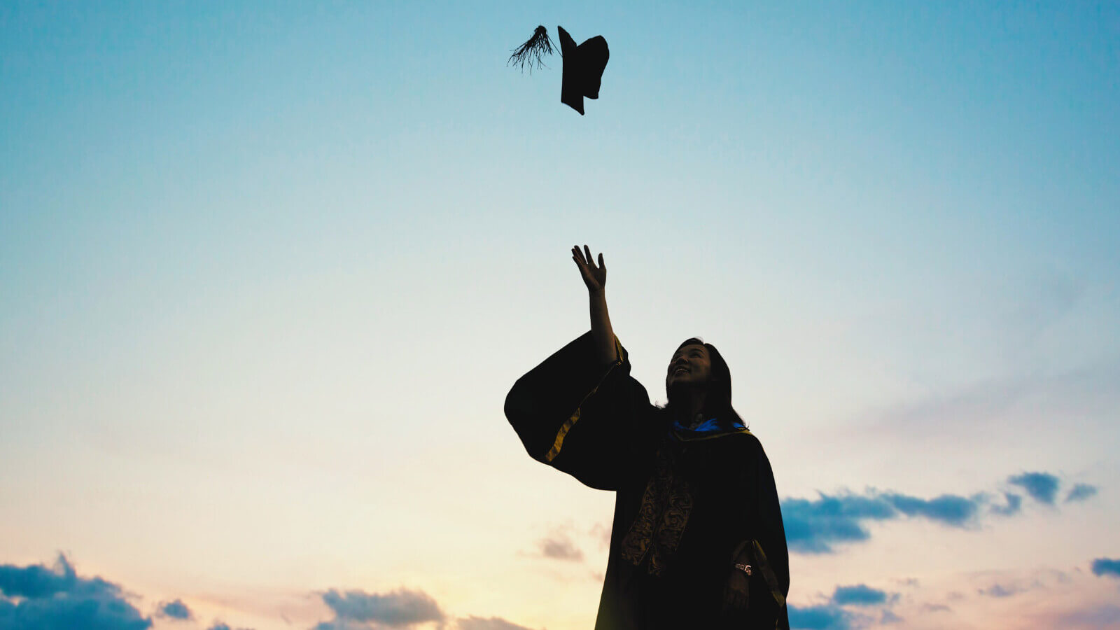 a student shadowed by the sunset, throws a graduation cap in the air