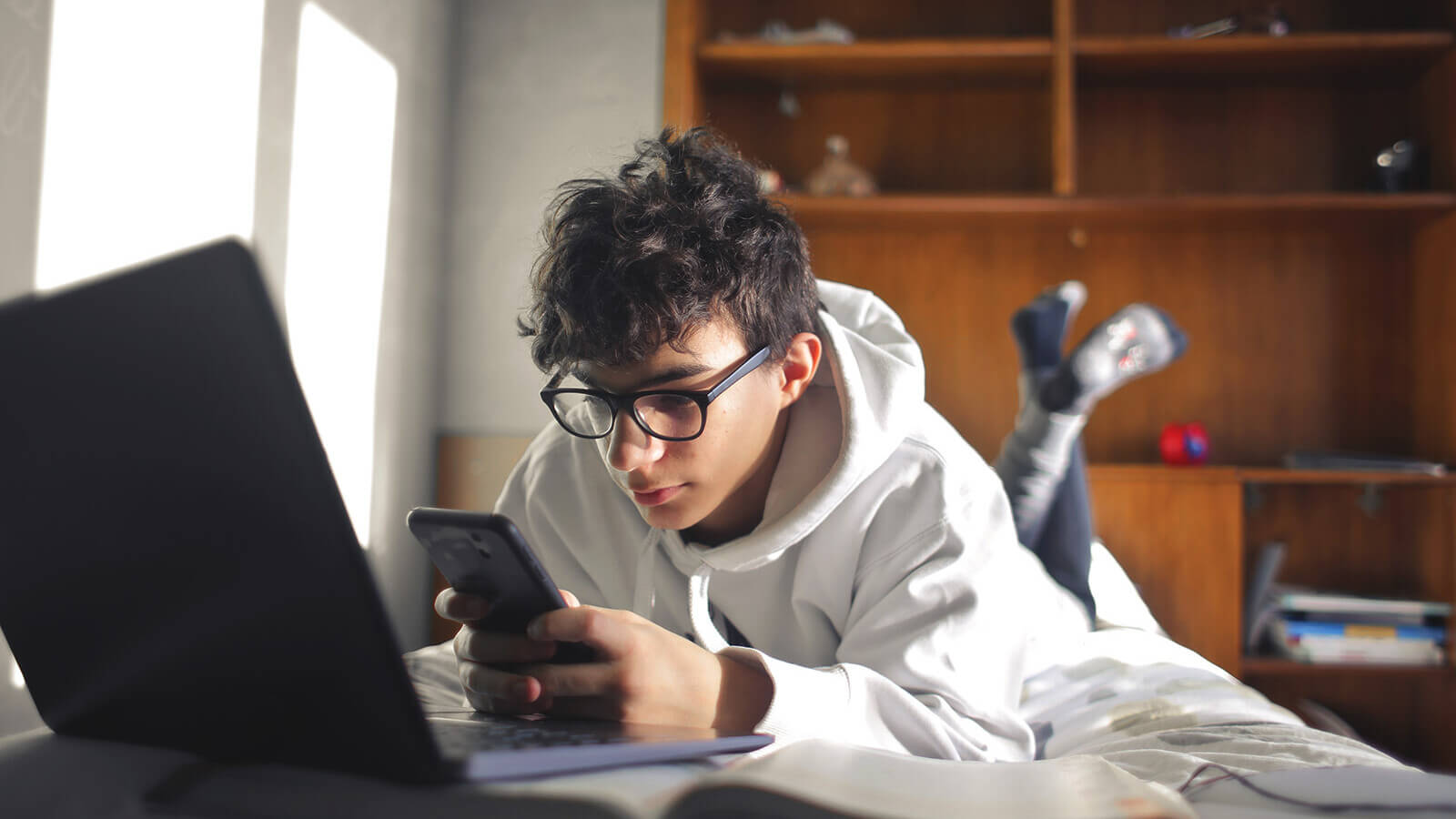 a student lays on his bed with his laptop open and looking at the phone in his had