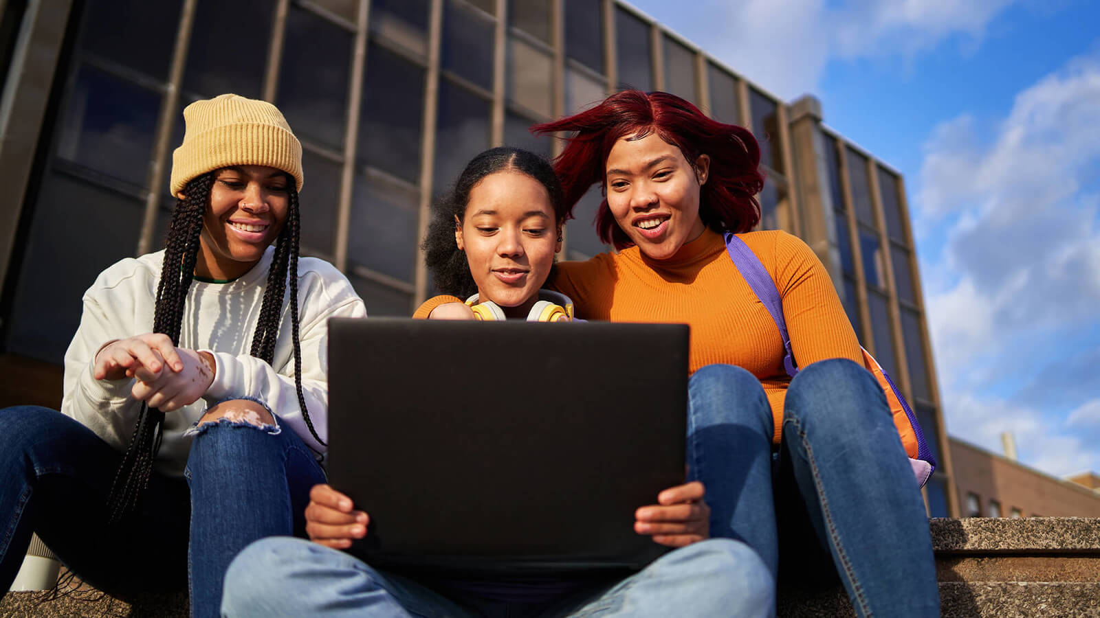 3 female students sitting outside on the steps looking at one black laptop