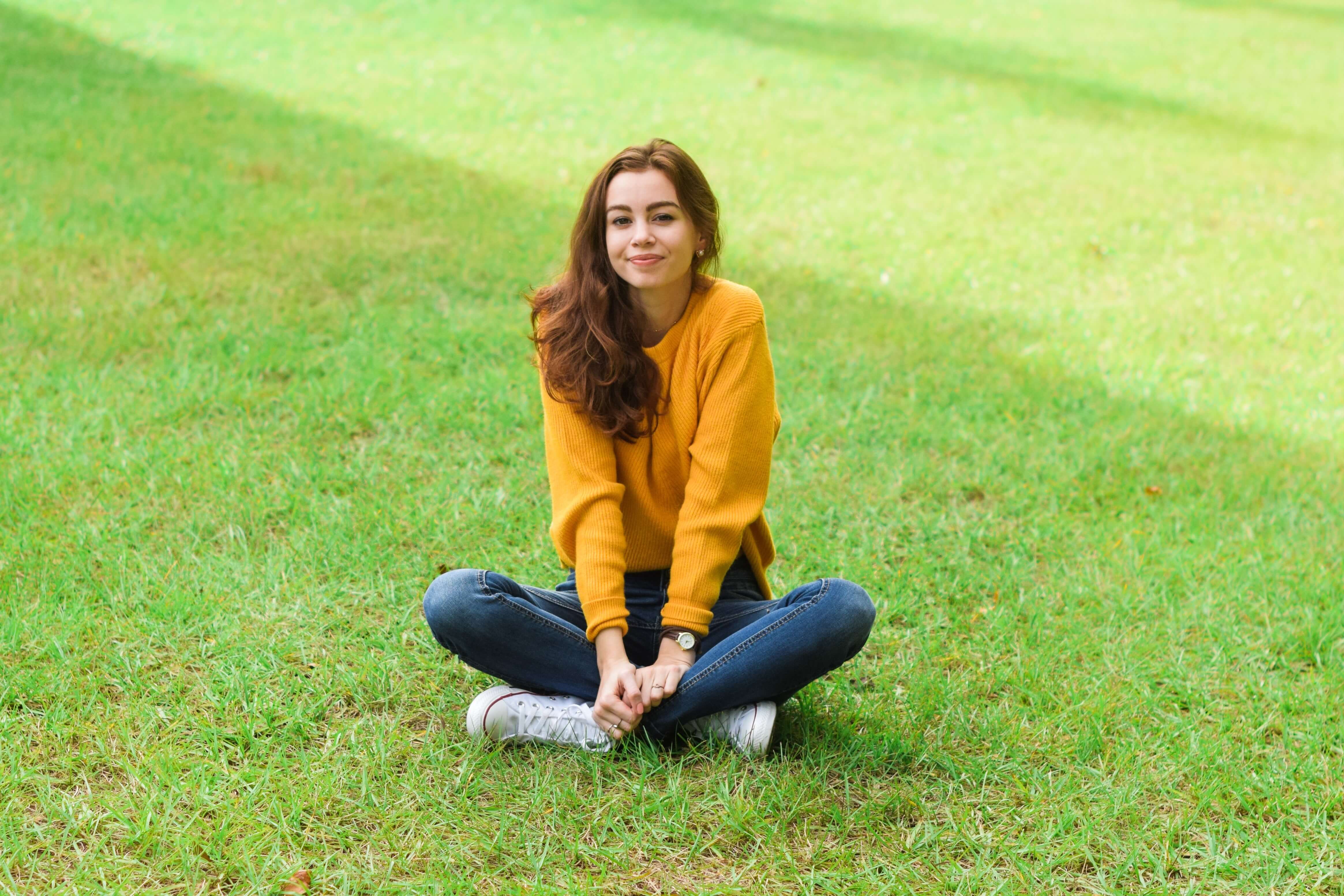 A young woman sitting cross-legged in the middle of a green lawn. She's wearing a mustard yellow sweater, blue jeans, and white sneakers. 