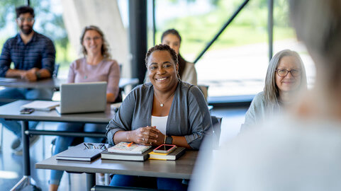 A group of people sitting at desks in a classroom setting, with a woman in focus smiling and listening attentively, while others around her take notes or use laptops.
