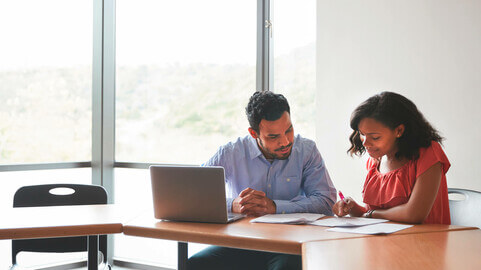 A teacher and student student sitting at a table with a laptop reviewing papers together.