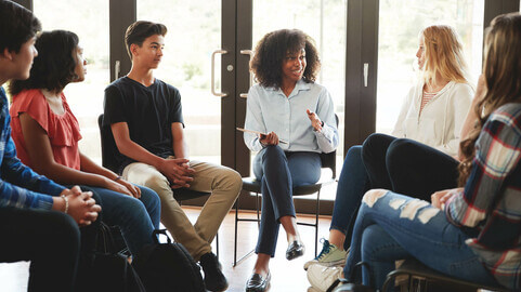 A woman holding a notebook sitting in a semi circle students as they engage in conversation.