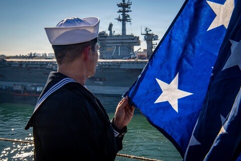 Navy service member holds the US flag as he watches a carrier go by