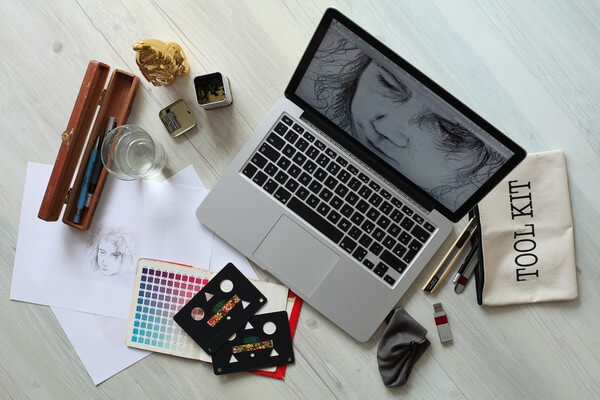 On a tabletop, a laptop is open with the screen showing a drawn face, likely male. Around the laptop are an array of drawing tools, including a cream-colored canvas pencil bag that says "TOOL KIT," a USB drive, a page of color swatches, tracing sheets for shapes, a few white pages of paper with the same sketched face that's on the laptop screen, a wooden case for drawing pens and pencils, a pencil sharpener, and a glass of water. 