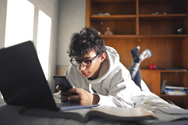 a student lays on his bed with his laptop open and looking at the phone in his had