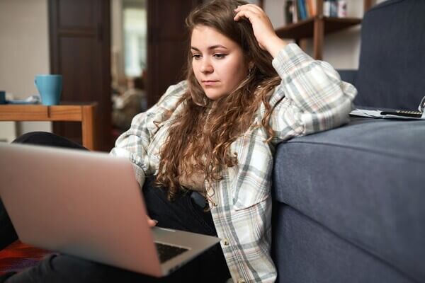 a student sits on the couch working on her laptop, looking stressed. 