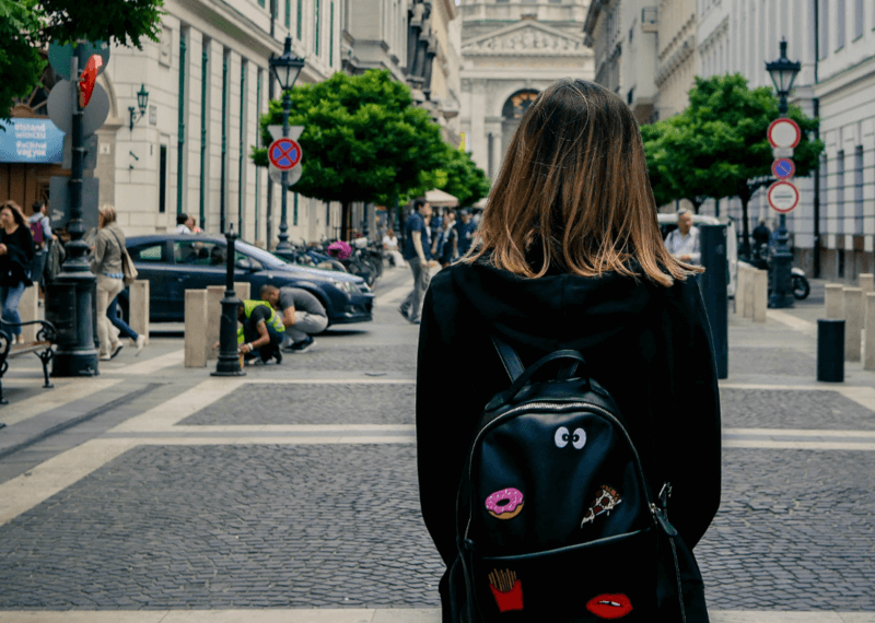 A young woman stands in the middle of a bustling city. She's wearing a backpack and has her back to the camera. 