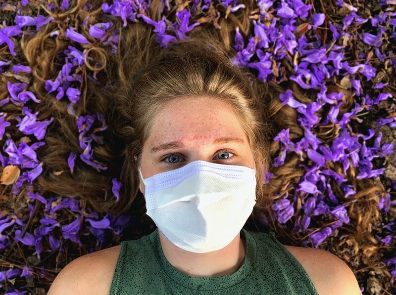 A young woman lays in a bed of purple flowers with her hair splayed out around her head. She is wearing a sleeveless, green top and a white mask over her face. 