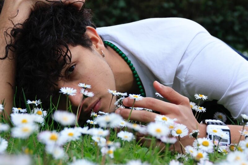 A young man is laying in the grass facing the camera. The grass is dotted with white flowers. 