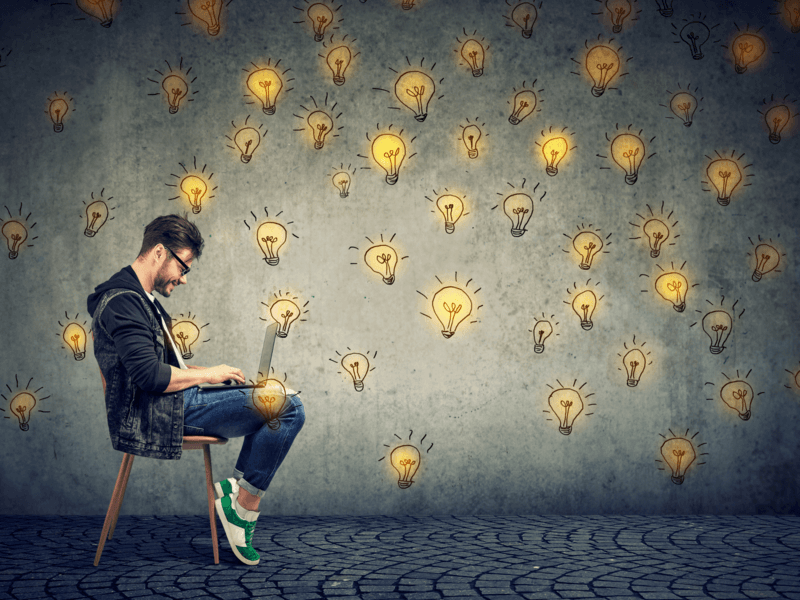 A man is sitting in a chair with a laptop on his legs. Drawn bright yellow lightbulbs surround him. 