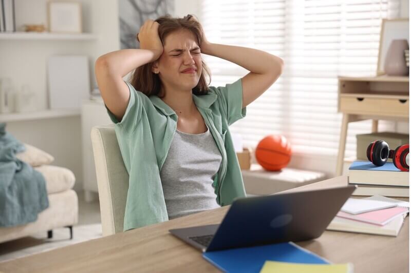 Stressed student sitting at desk grabbing head and staring at laptop trying to pick her right-fit college
