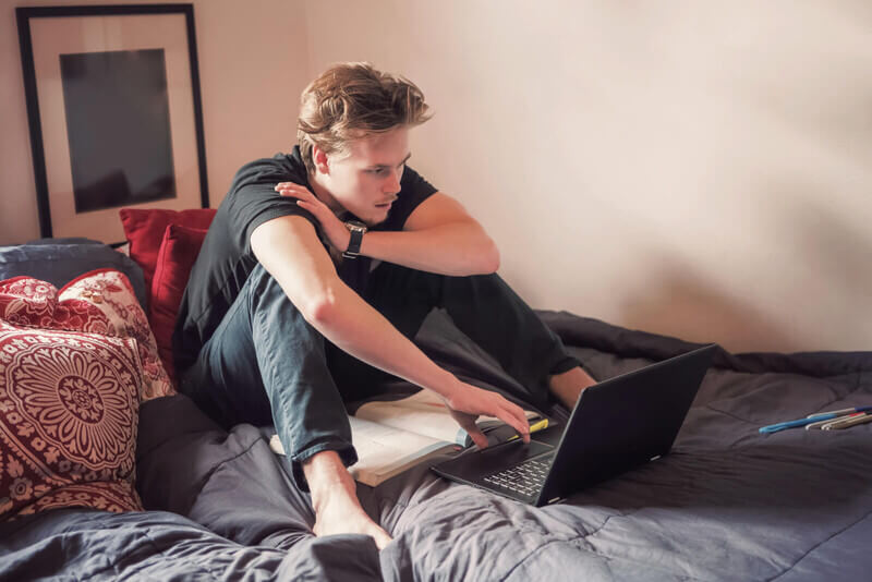 a student sits on a bed and does class work on a laptop