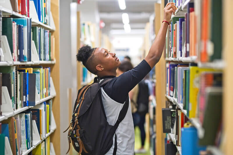 A male student in the library reaching the top shelf.